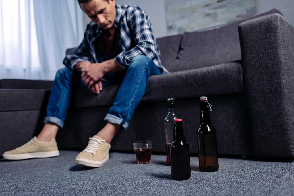 мужчина смотрит на алкоголь на полу, сидя дома на диване
 - Фото, изображение