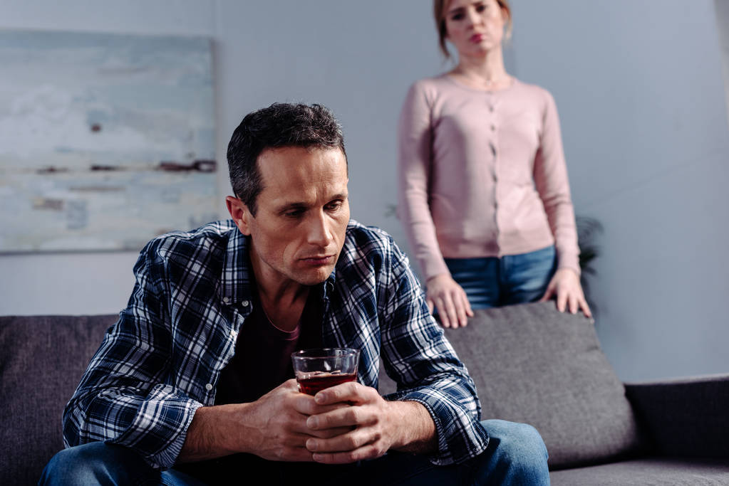 homem com copo de álcool sentado no sofá, enquanto esposa de pé atrás em casa
 - Foto, Imagem
