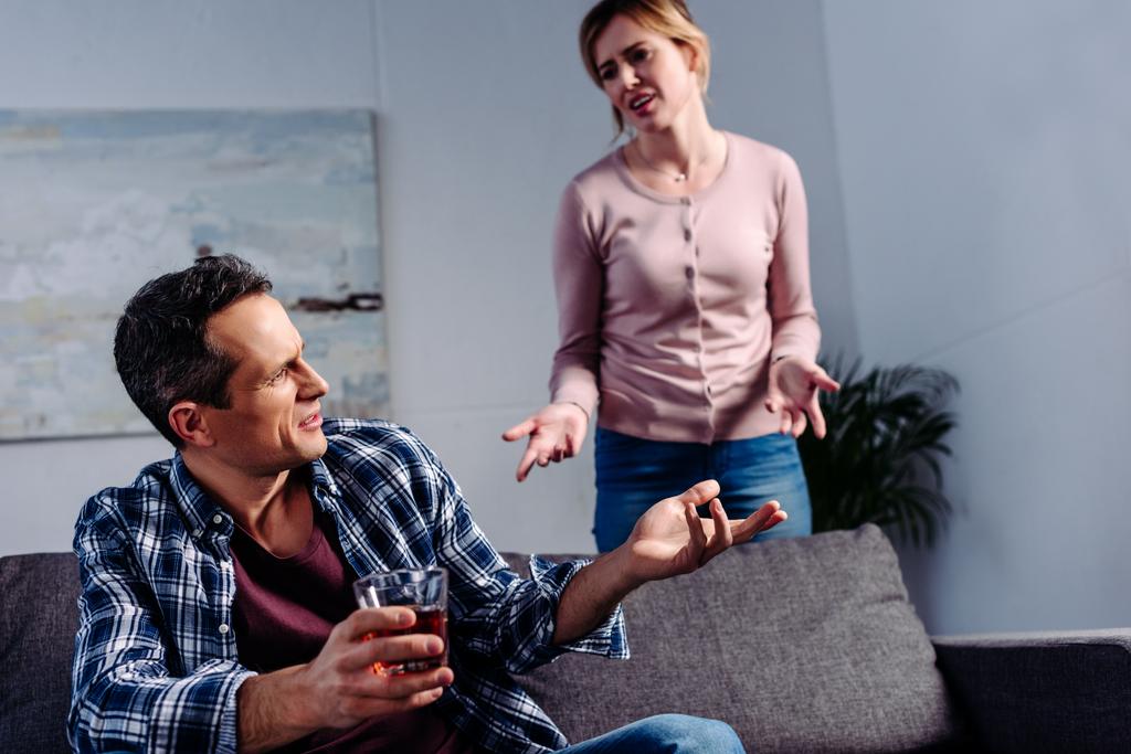 чоловік зі склянкою алкоголю сидить на дивані, сперечаючись з дружиною позаду вдома
 - Фото, зображення