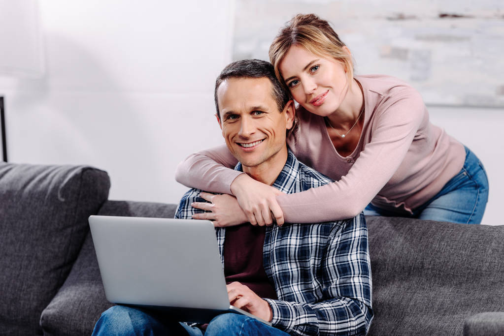 portrait of woman hugging husband with laptop on sofa at home - Photo, Image