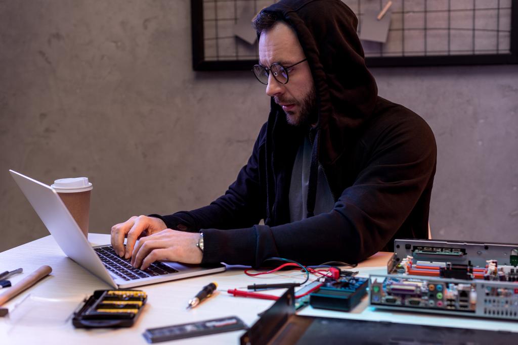 ο άνθρωπος σε hoodie χρησιμοποιώντας φορητό υπολογιστή κοντά σε σπασμένα pc  - Φωτογραφία, εικόνα