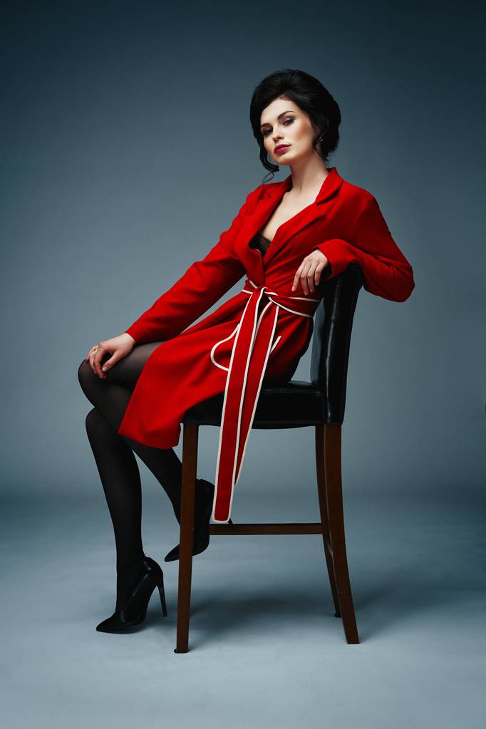 Красивая молодая женщина в стильном пальто сидит на стуле на фоне студии
  - Фото, изображение