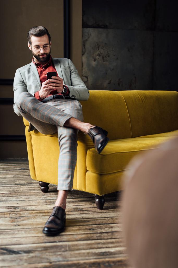 bello uomo elegante utilizzando smartphone e seduto sul divano giallo
  - Foto, immagini