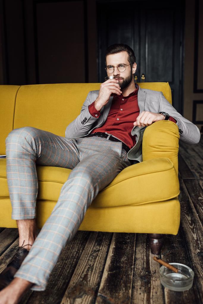 όμορφος άνδρας με ποτήρι ουίσκι που κάθεται στον καναπέ με πούρο σε τασάκι στο πάτωμα  - Φωτογραφία, εικόνα