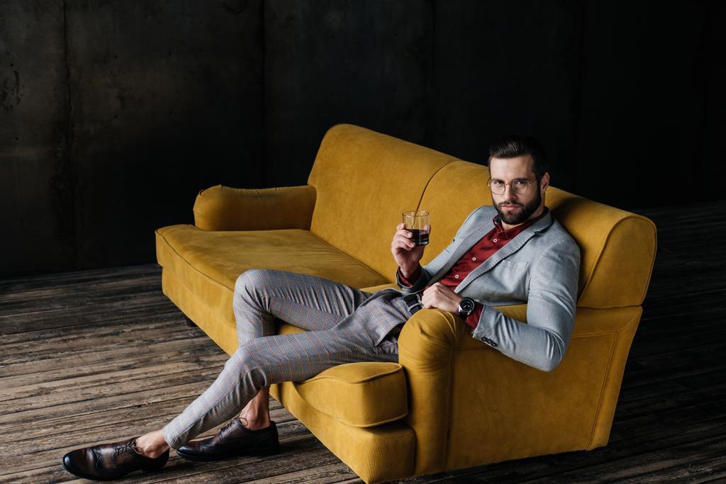 κομψό όμορφος άνδρας κρατά ποτήρι κονιάκ και κάθεται στον καναπέ κίτρινο - Φωτογραφία, εικόνα