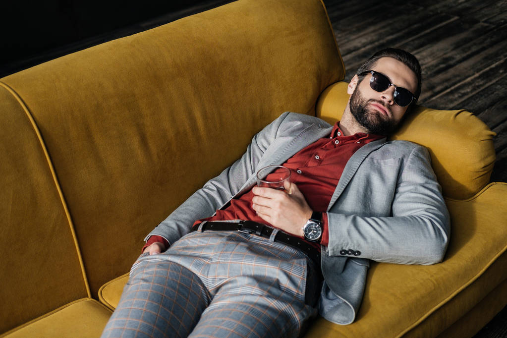 κομψό μεθυσμένος άντρας με γυαλιά ηλίου με ποτηράκι κονιάκ ξαπλωμένη στον καναπέ κίτρινο - Φωτογραφία, εικόνα