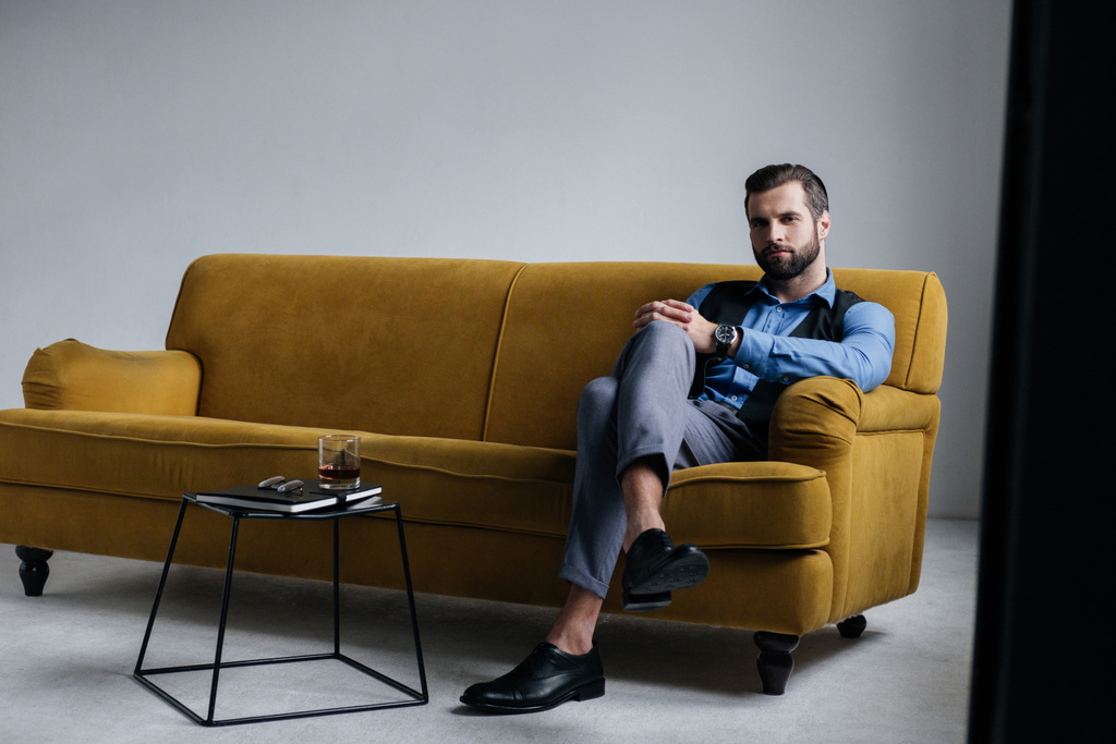 модный мужчина сидит на желтом диване с алкогольным напитком на столе рядом
 - Фото, изображение