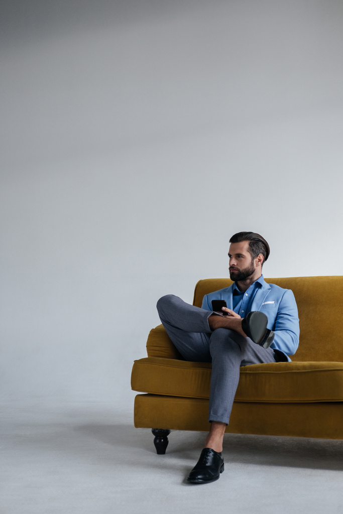stylish man in blue trendy suit holding smartphone and sitting on yellow sofa - Photo, Image