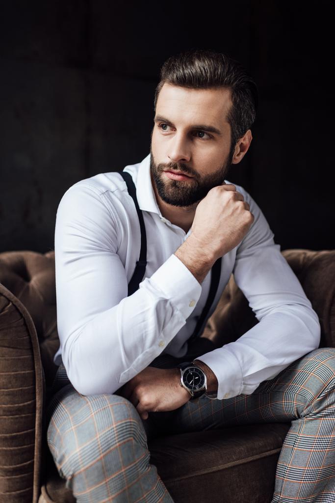stijlvolle man in bretels denken en zitten in een leunstoel  - Foto, afbeelding