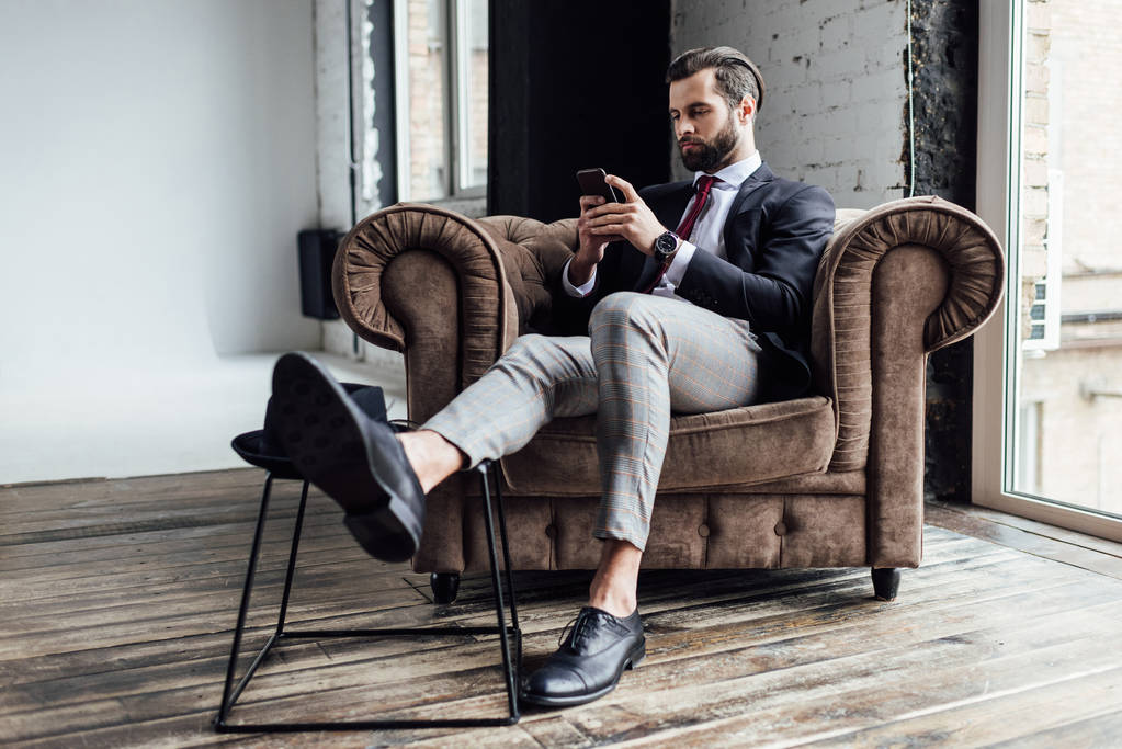 stylish businessman using smartphone and sitting in armchair in loft - Photo, Image