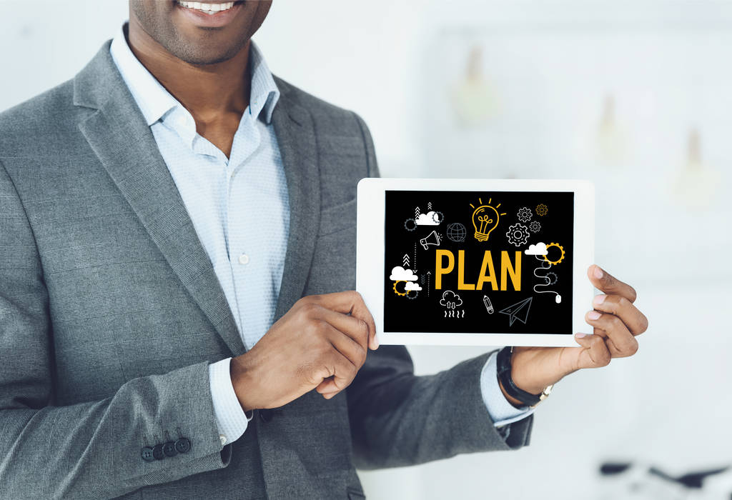 обрезанное изображение улыбающегося африканского американца, показывающего цифровой планшет со словом "план" и бизнес-иконками
 - Фото, изображение