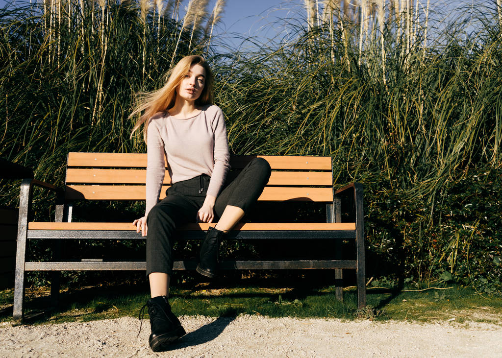 ładny piękny blond dziewczyna siedzi na ławce w przyrodzie w słońcu, jej włosy trzepotanie - Zdjęcie, obraz