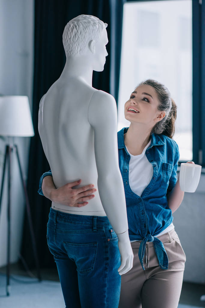 Zijaanzicht van gelukkige vrouw met kopje koffie knuffelen mannequin, unidirectioneel love concept - Foto, afbeelding