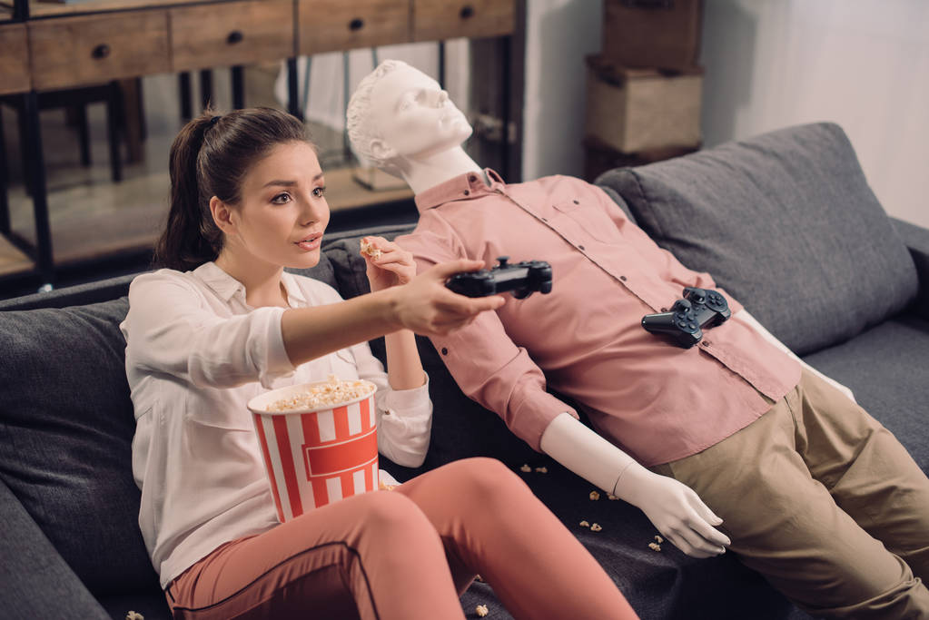 Młoda kobieta jedzenie popcorn, grając w gry wideo z manekina w pobliżu przez w domu, pojęcie samotności - Zdjęcie, obraz