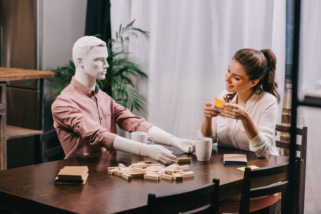 jovem mulher jogando jogo junto com boneca leigo à mesa com xícaras de café, conceito de solidão
 - Foto, Imagem