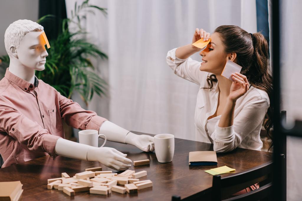 jonge vrouw speelspel samen met leek pop aan tafel met kopjes koffie, eenzaamheid concept - Foto, afbeelding