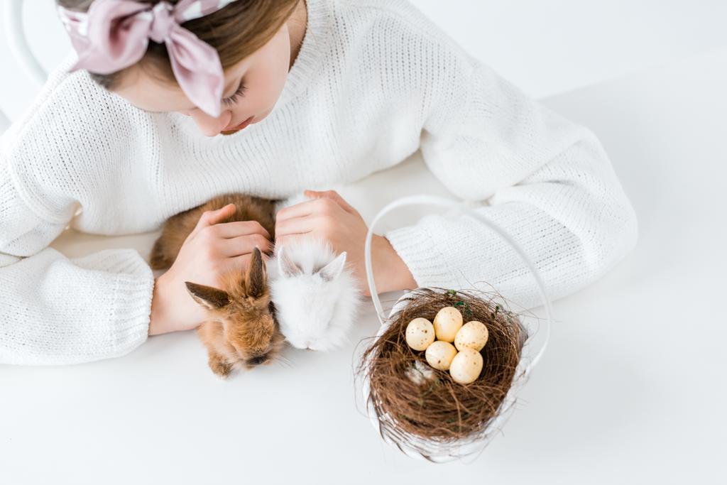 обрізаний знімок дівчини, дивлячись на милих пухнастих кроликів і кошик з великодніми яйцями в гнізді
  - Фото, зображення