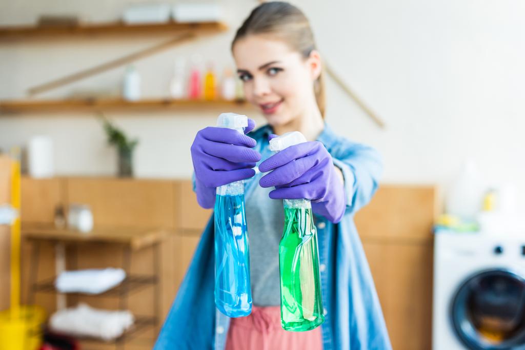 jovem mulher em luvas de borracha segurando garrafas de plástico com fluidos de limpeza e sorrindo para a câmera
  - Foto, Imagem