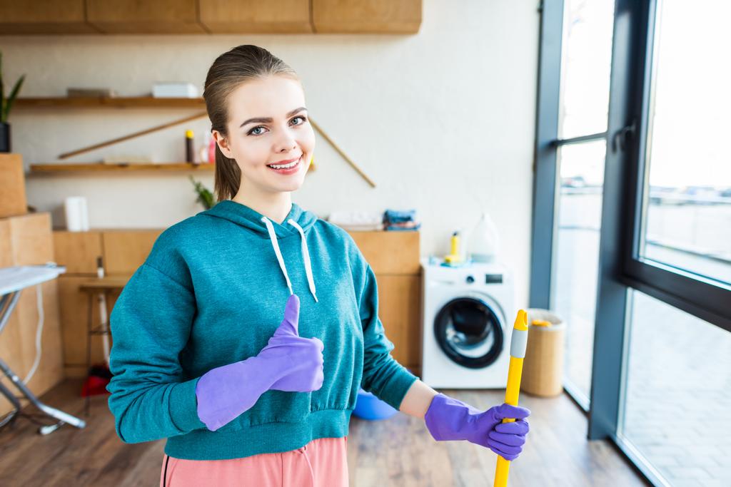 Lächelnde junge Frau putzt Haus mit Wischmopp und zeigt Daumen hoch - Foto, Bild