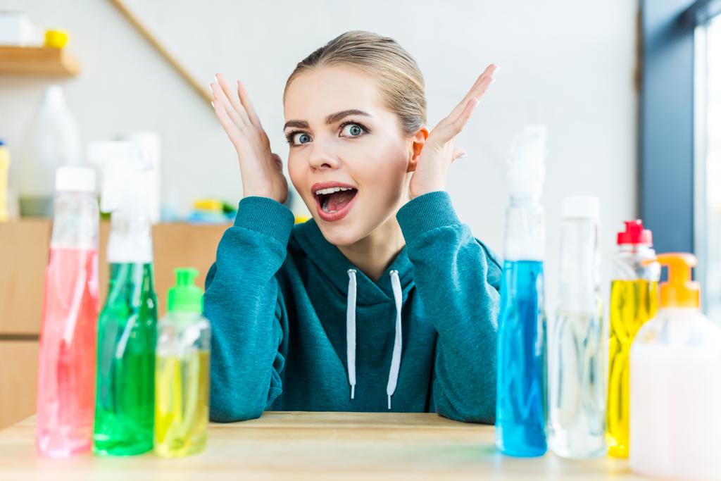 σοκαρισμένος γυναίκα βλέπει την κάμερα ενώ κάθεται στο τραπέζι με πλαστικά μπουκάλια με προϊόντα καθαρισμού - Φωτογραφία, εικόνα