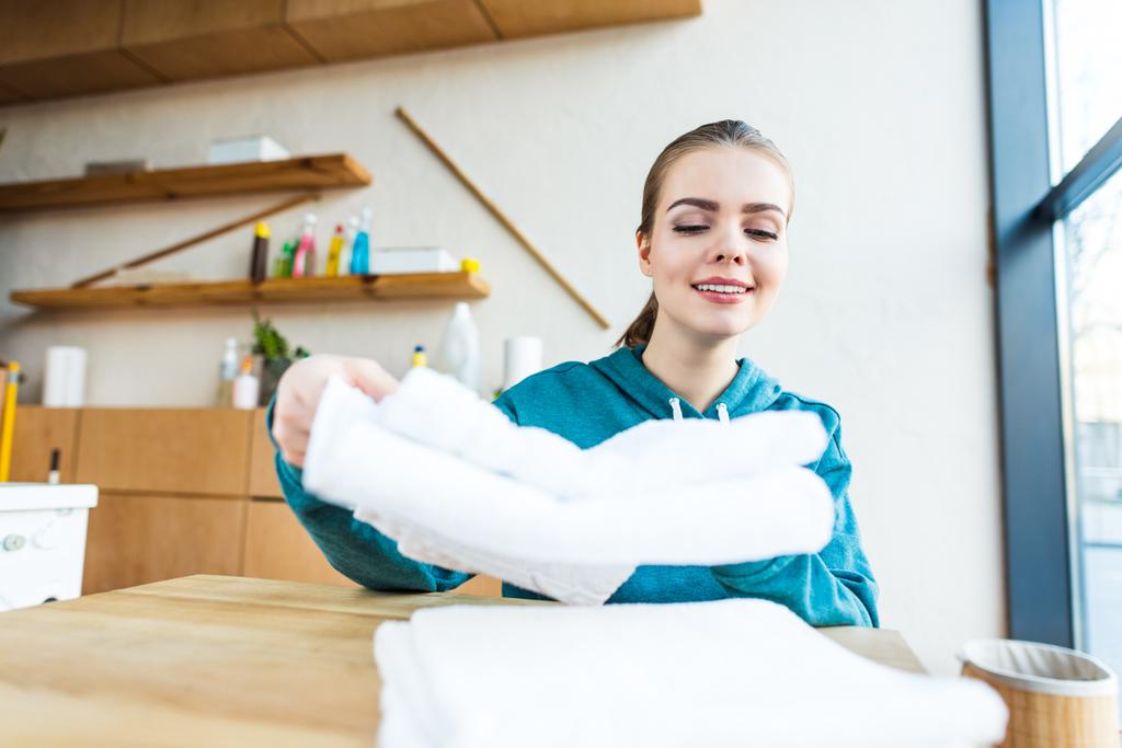 Lächelnde junge Frau stapelt saubere weiße Handtücher auf dem Tisch - Foto, Bild
