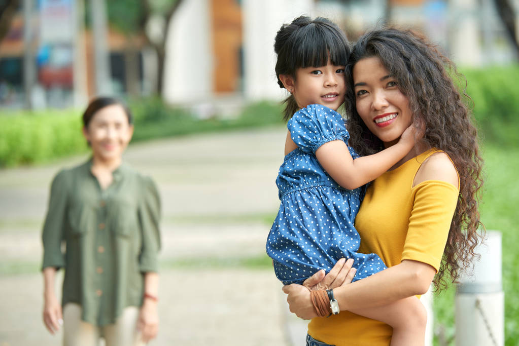 Sevimli küçük kızını silah, yeşil Park arka plan üzerinde tutan çekici Asyalı kadın - Fotoğraf, Görsel