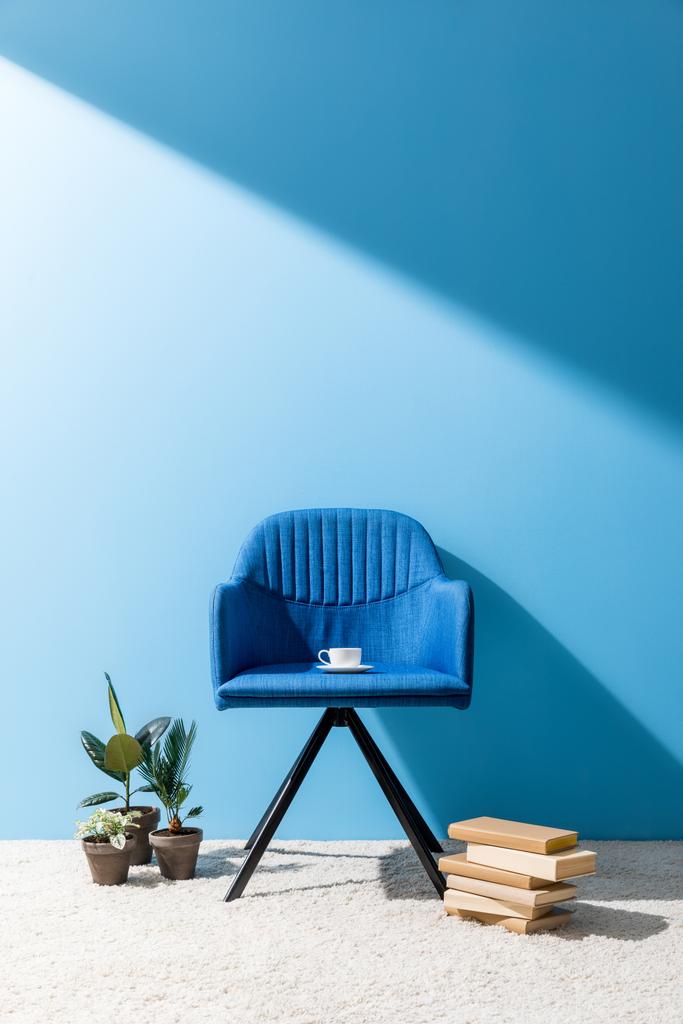 blauer Sessel mit Tasse Kaffee und Büchern mit Blumentöpfen auf dem Boden vor blauer Wand - Foto, Bild