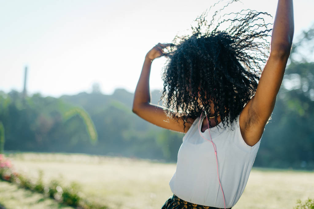 Attiva giovane bella ragazza africana sta ballando emotivamente e scuotendo i suoi capelli ricci scuri mentre ascolta musica con le cuffie sul prato soleggiato
. - Foto, immagini