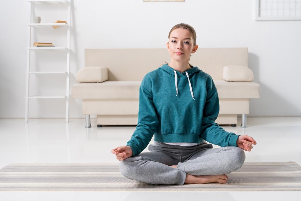 chica sentada en posición de loto en estera de yoga en casa y mirando a la cámara
 - Foto, Imagen