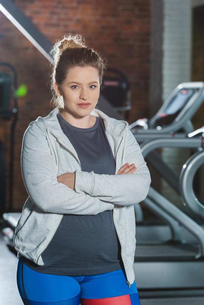 Übergewichtige Frau steht mit verschränkten Armen im Fitnessstudio und blickt in die Kamera - Foto, Bild