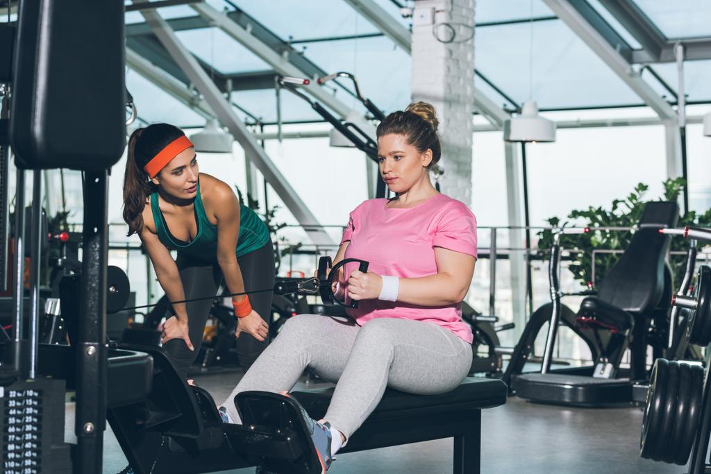 vrouw uit te werken op training apparaat terwijl trainer kijken naar haar overgewicht  - Foto, afbeelding