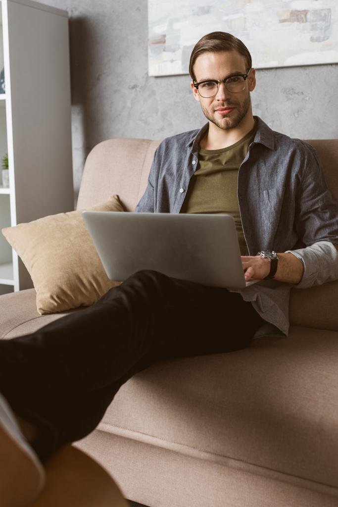 стильный человек работает с ноутбуком на диване и смотрит в камеру
 - Фото, изображение
