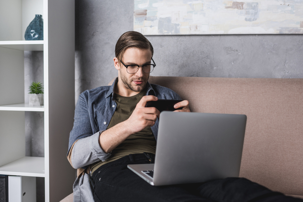молодой фрилансер с ноутбуком на коленях сидит на диване и играет в игру на смартфоне
 - Фото, изображение