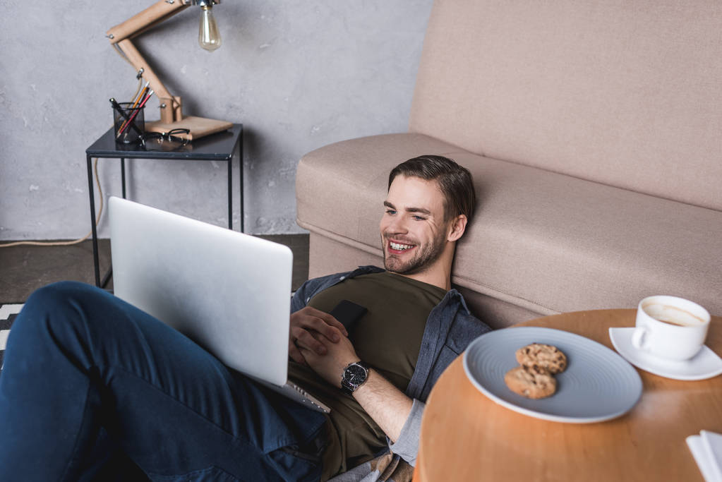 молодий щасливий чоловік використовує ноутбук, сидячи на підлозі
 - Фото, зображення