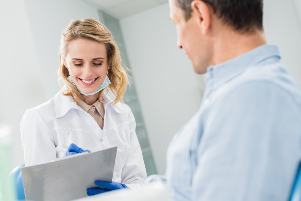 Patient and smiling doctor recording diagnosis in modern dental clinic - Photo, Image