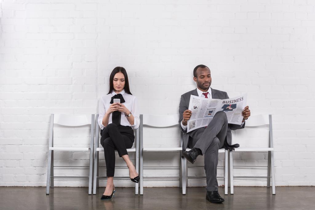 Aziatische zakenvrouw met smartphone en Afro-Amerikaanse zakenman met krant wachten op sollicitatiegesprek  - Foto, afbeelding