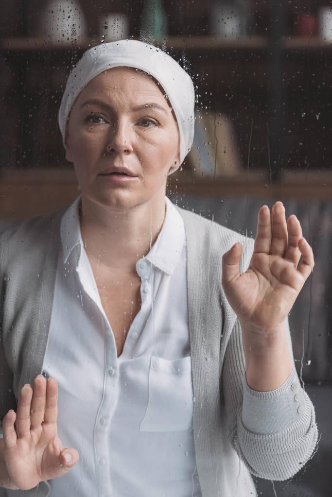 malade femme mûre en mouchoir regardant caméra à travers le verre avec des gouttes de pluie
 - Photo, image
