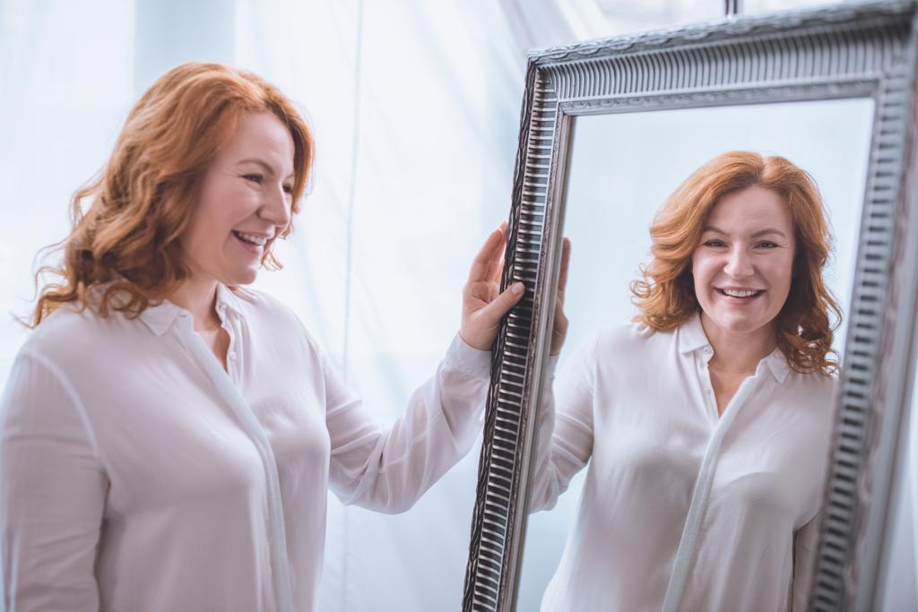 mooie lachende volwassen vrouw staande in de buurt van spiegel en reflectie kijken - Foto, afbeelding