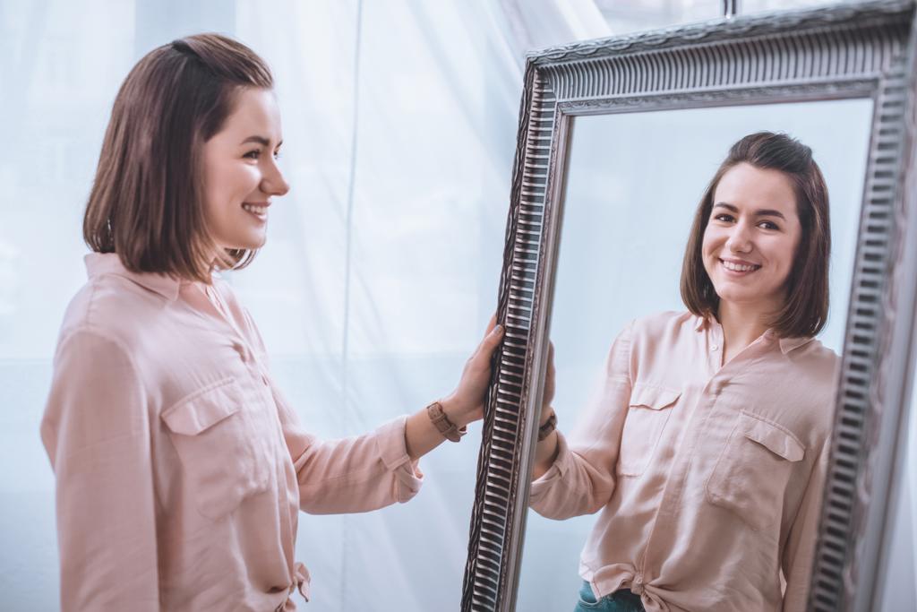 mooie jonge vrouw staande in de buurt van spiegel en reflectie kijken glimlachen - Foto, afbeelding