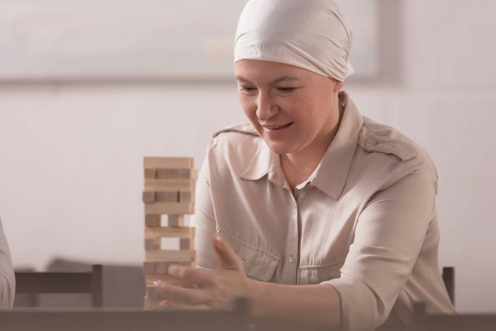 sick smiling mature woman in kerchief building tower from wooden blocks at home - Photo, Image