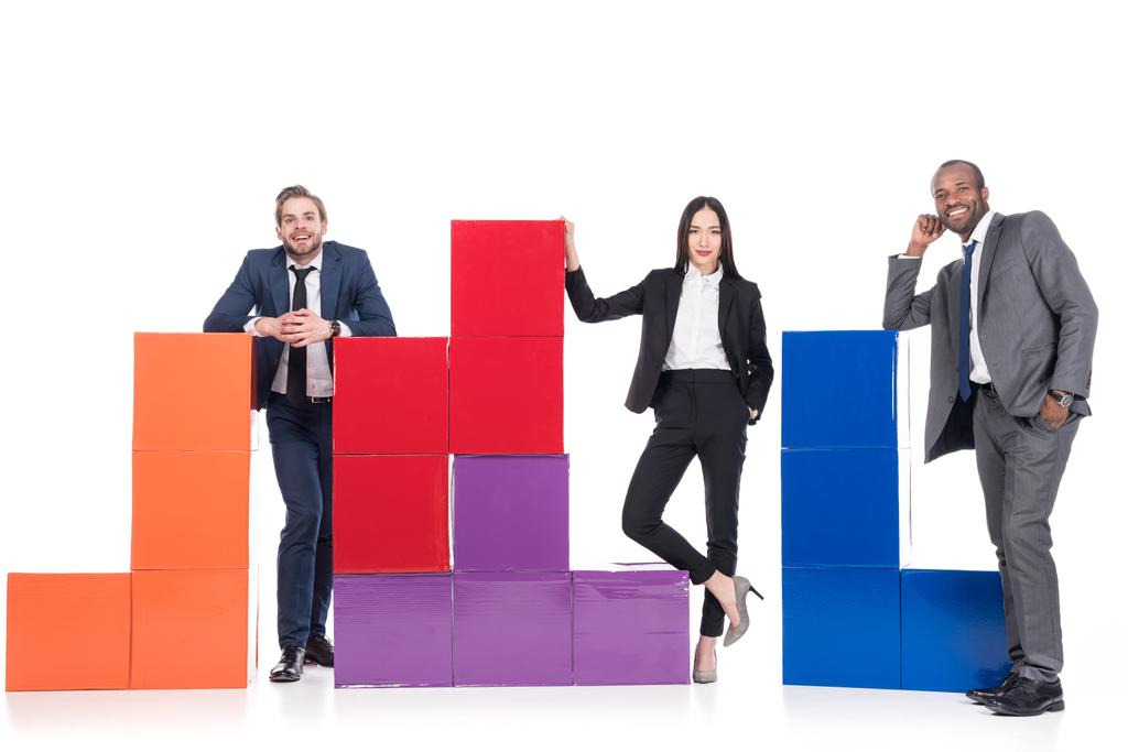 улыбающиеся многонациональные бизнесмены, стоящие рядом с красочными блоками, изолированными по белой концепции командной работы
 - Фото, изображение