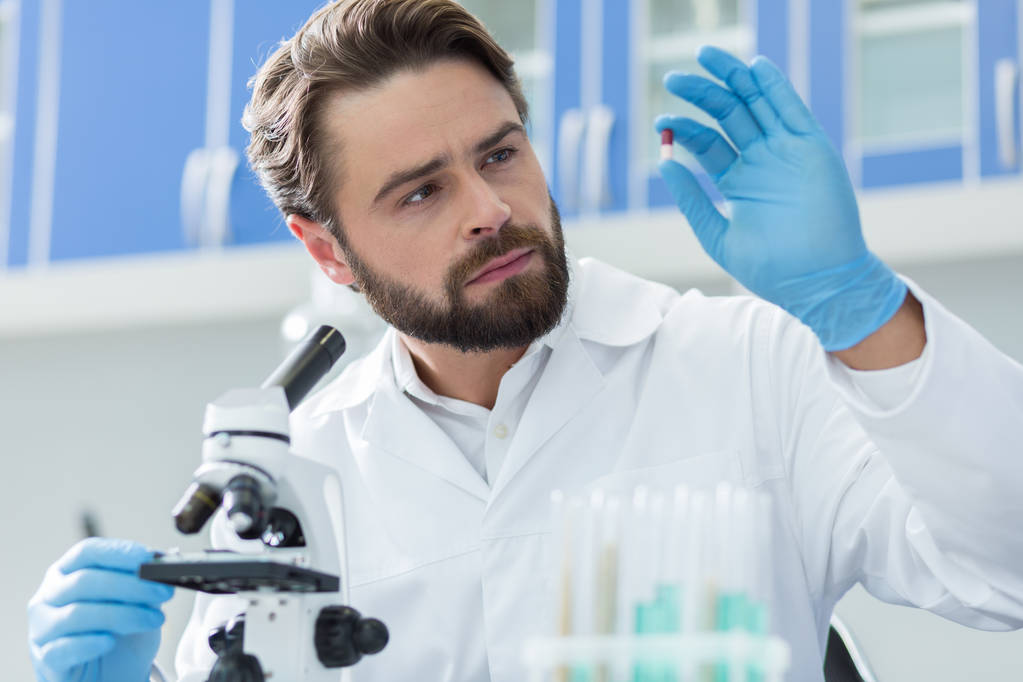 Yenilikçi tedavi. Akıllı yakışıklı güzel bilim adamı mikroskop önünde oturan ve yeni ilaç geliştirirken hap bakarak - Fotoğraf, Görsel