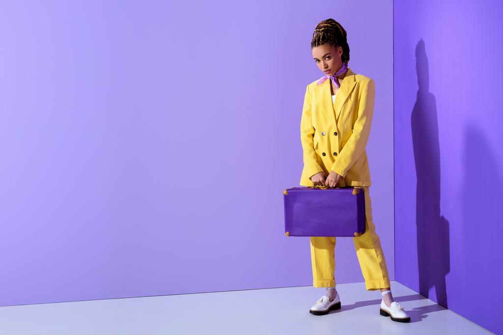紫のスーツケースとトレンディな黄色いスーツでポーズをとってアフリカ系アメリカ人の女の子 - 写真・画像