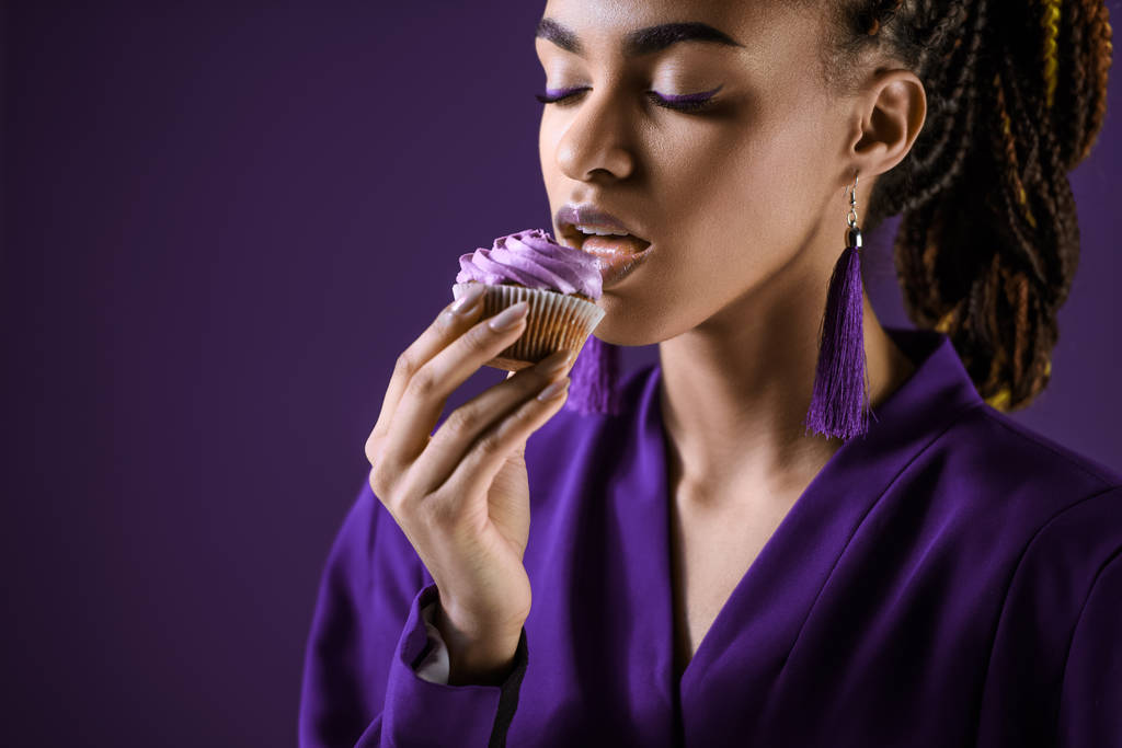 Πάθος αφρικανικό αμερικανικό κορίτσι σε μωβ σακάκι τρώει cupcake, απομονώνονται σε μωβ - Φωτογραφία, εικόνα