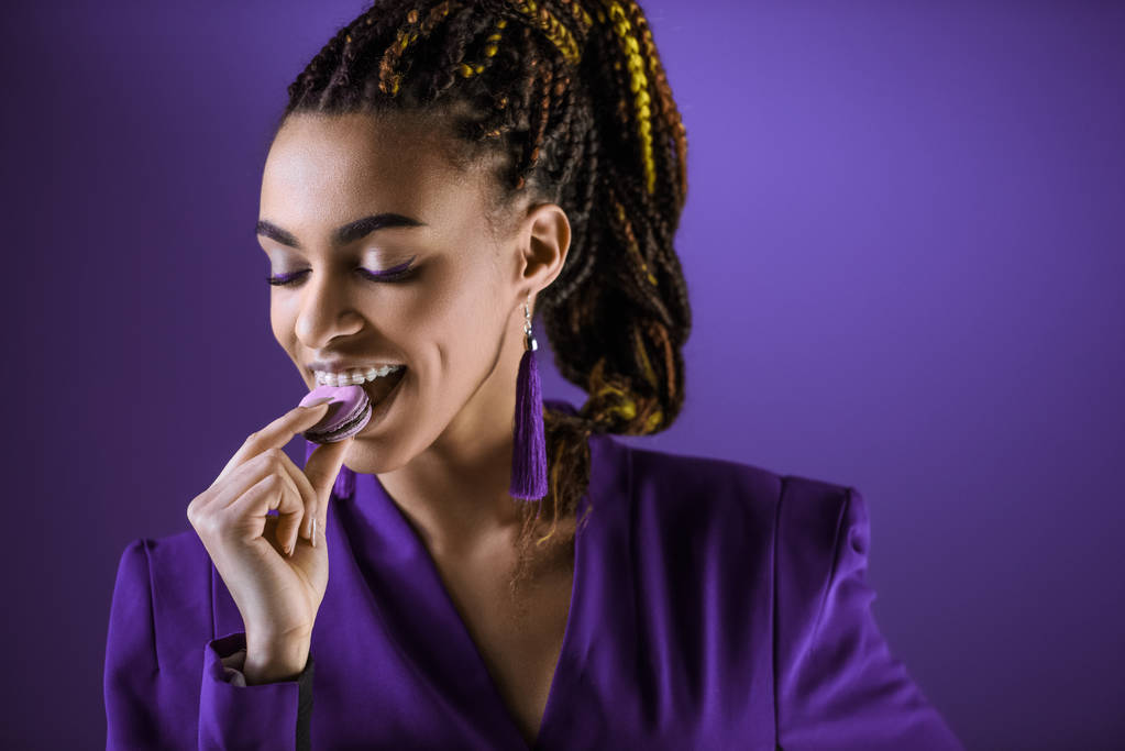 веселая девушка мулатка ест макарон, изолированный на фиолетовый
 - Фото, изображение