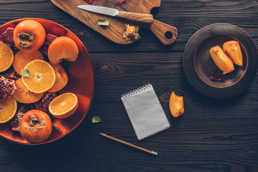 top view of persimmons, oranges and pomegranates with notebook on table - Photo, Image