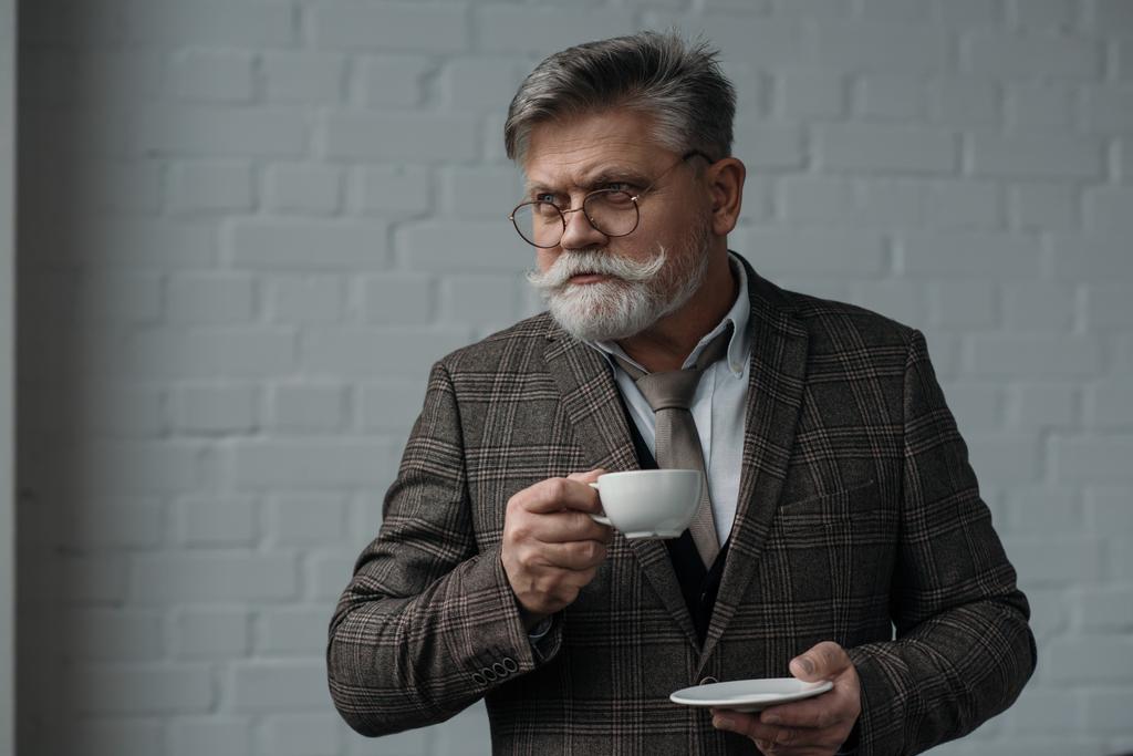 白いレンガの壁の前にコーヒーのカップを持つハンサムな年配の男性 - 写真・画像