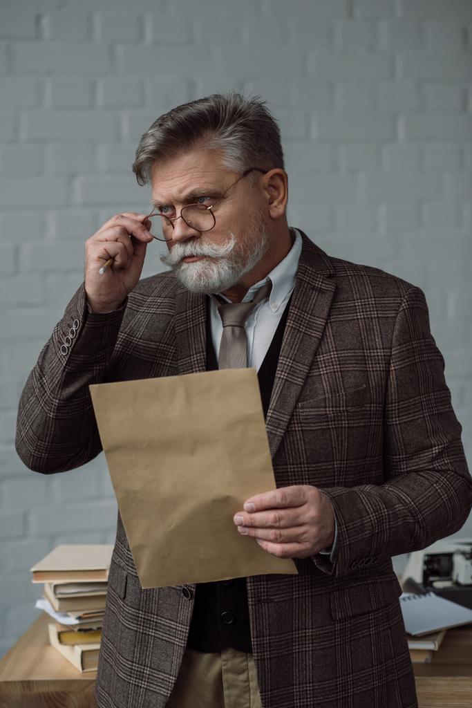 senior schrijver in tweed pak en brillen met letter in de buurt van werkplek - Foto, afbeelding