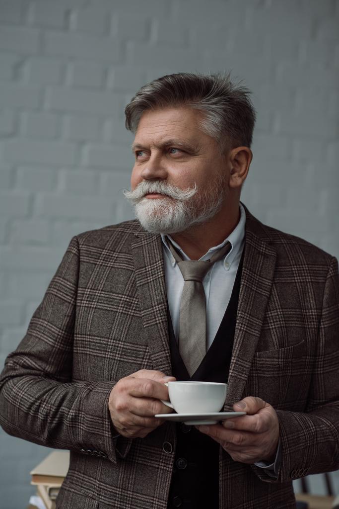 白いレンガの壁の前にコーヒーのカップを持つ年配の男性 - 写真・画像