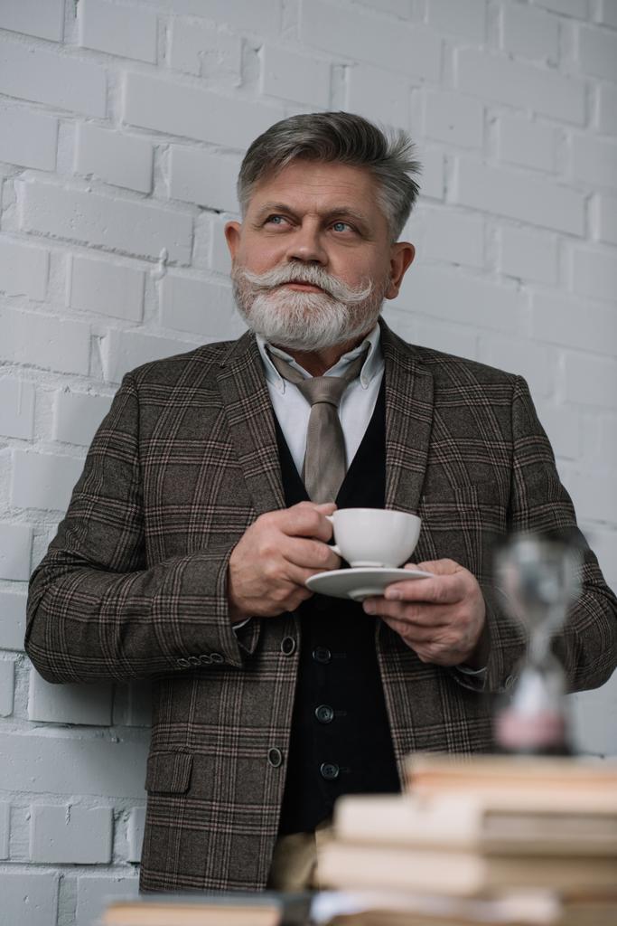 doordachte senior man drinken koffie met stapel boeken en zandloper op voorgrond - Foto, afbeelding